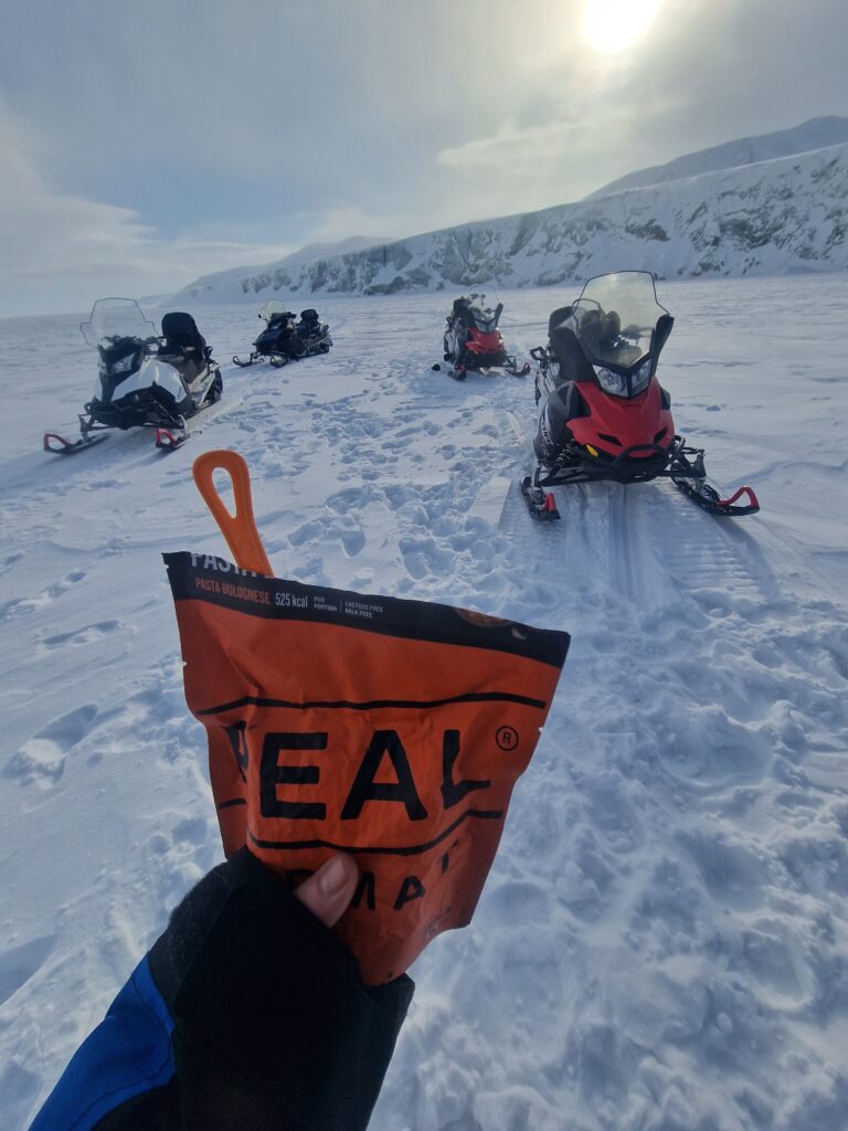 Alcune motoslitte davanti ad un ghiacciaio e una busta arancione di cibo liofilizzato